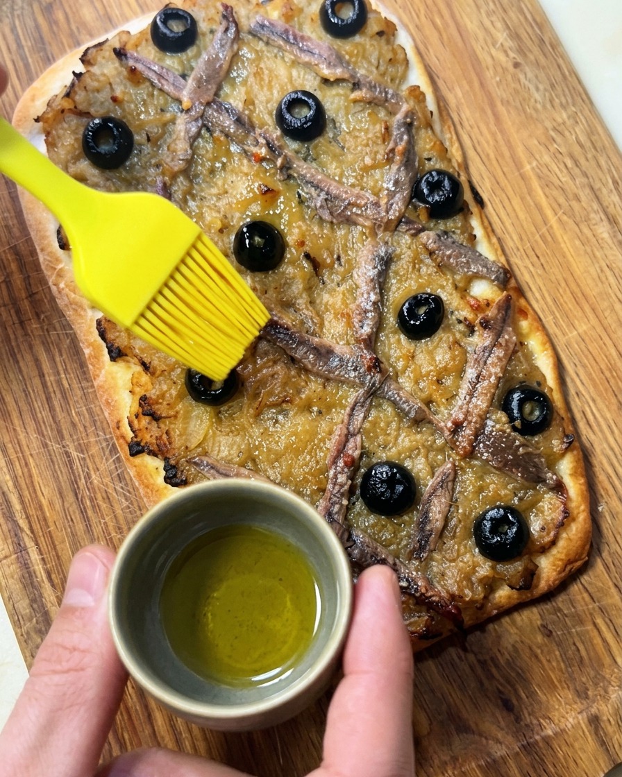 A hand using a yellow silicone brush to apply olive oil onto a freshly baked Pissaladière topped with onions, anchovies, and black olives.