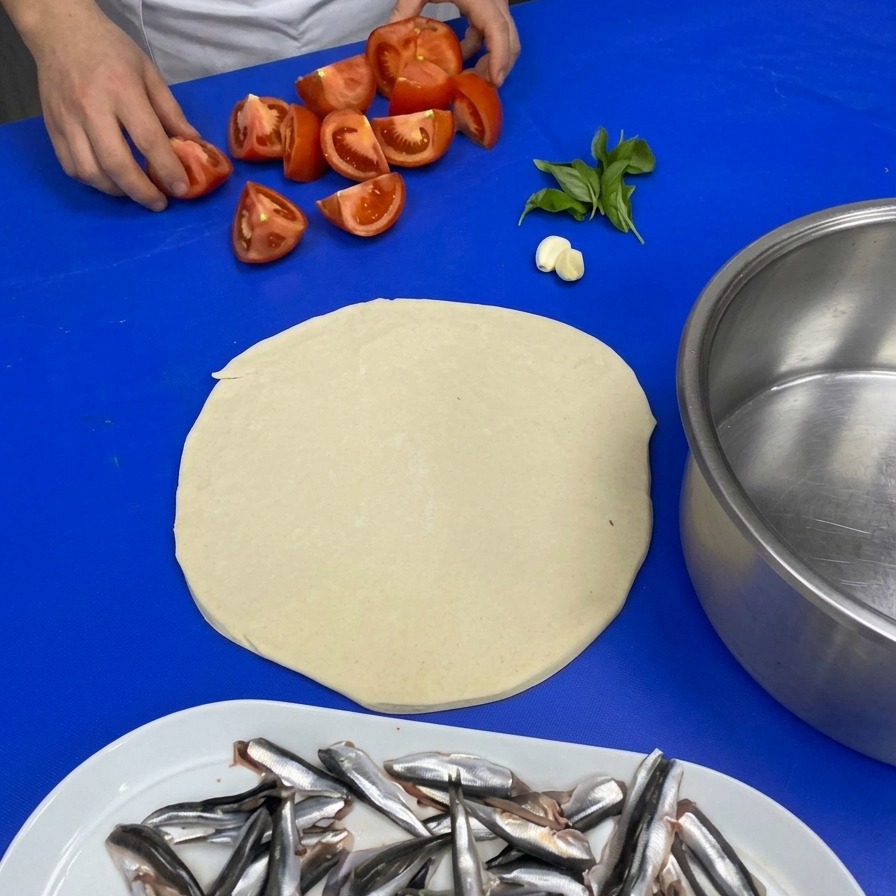 Chefs hands picking up quartered tomatoes from a blue work surface alongside a garlic clove and fresh basil leaves.