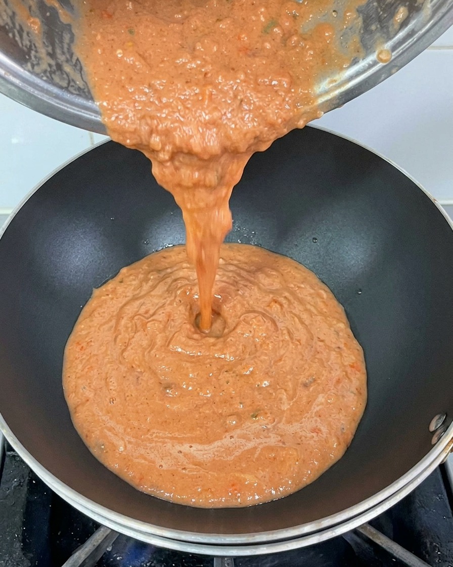 Pouring thick, blended red tomato sauce from a metal pot into a hot black wok.