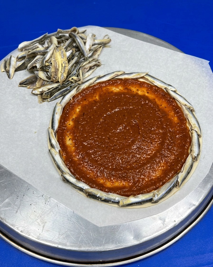 A perfectly round pizza base framed by a border of raw anchovies, with a smoothly spread layer of red tomato sauce covering the center.