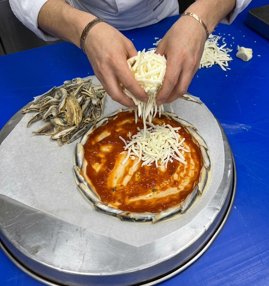 Hands generously sprinkling freshly grated white cheese over a pizza base coated with red tomato sauce and an anchovy crust.