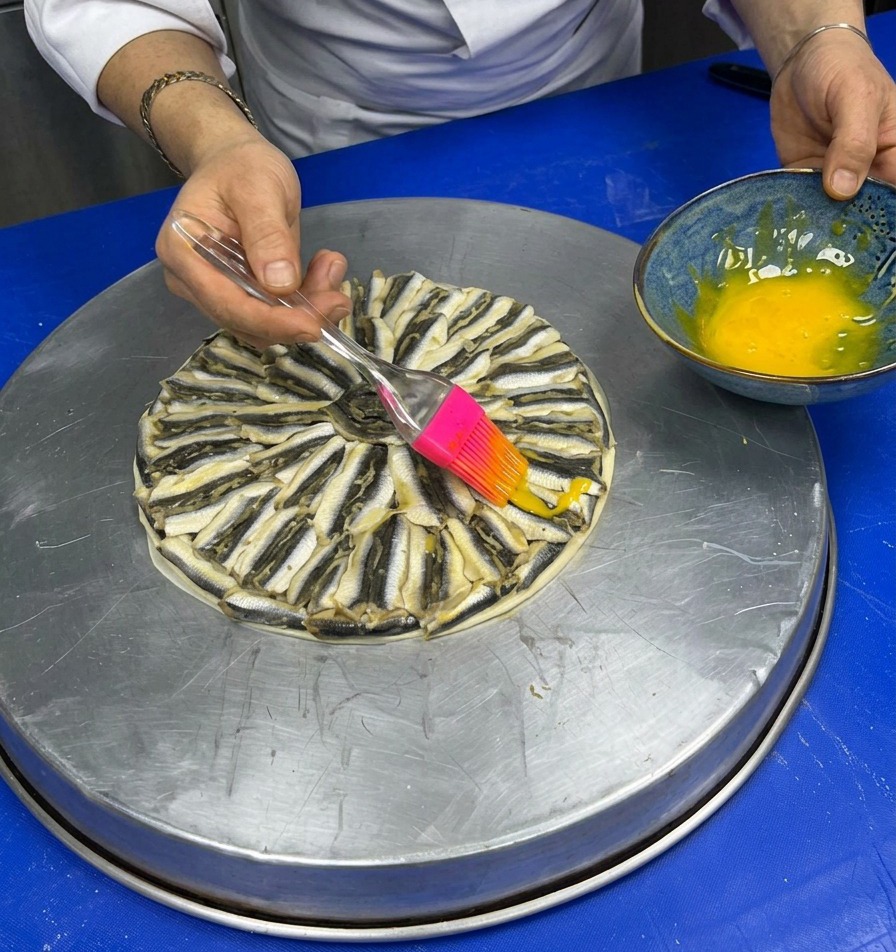 Using a pink silicone brush to lightly paint a yellow egg wash over a pizza completely topped with a circular arrangement of anchovies.