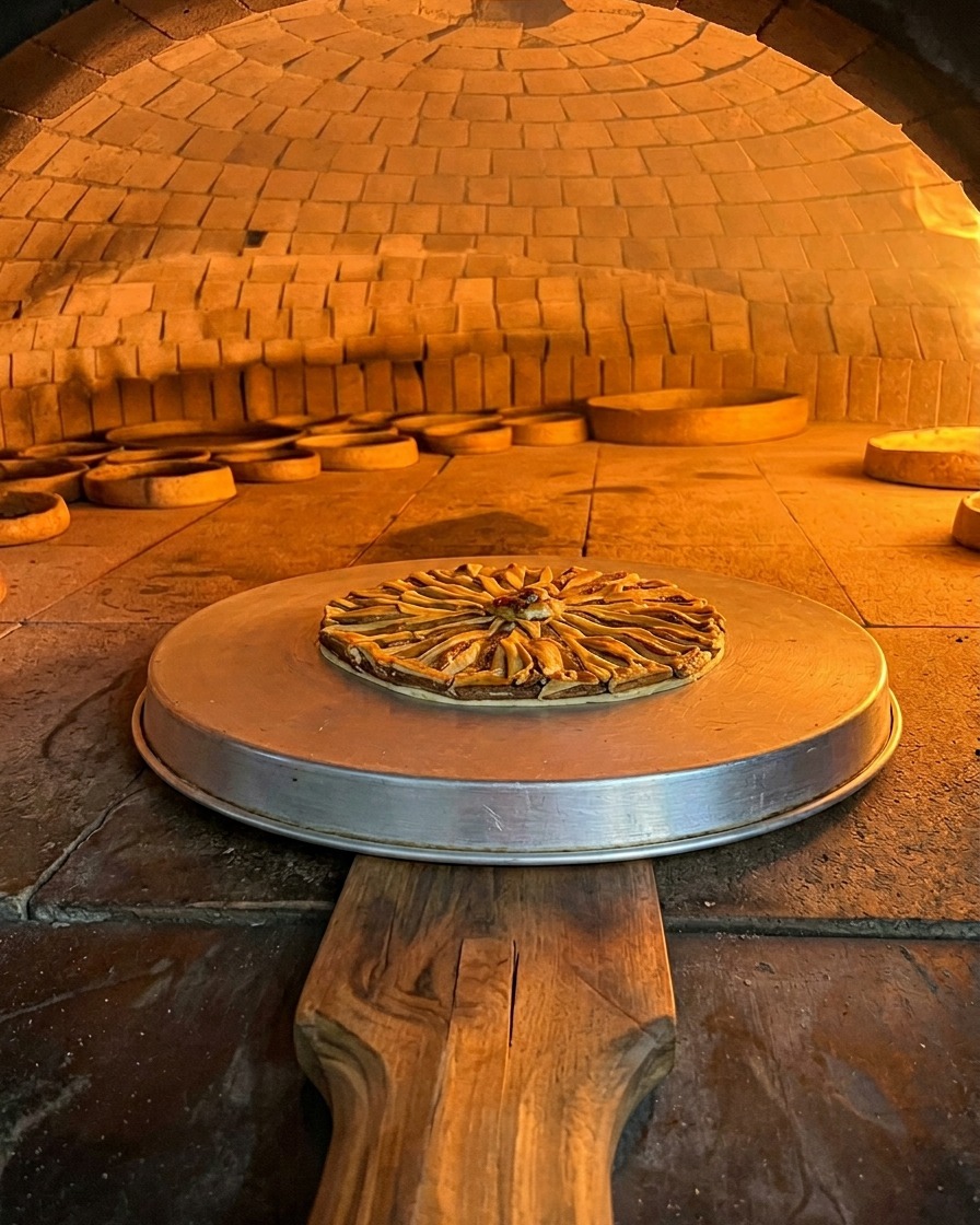 An anchovy-topped pizza sitting on a round metal tray baking inside the glowing orange chamber of a wood-fired brick oven.