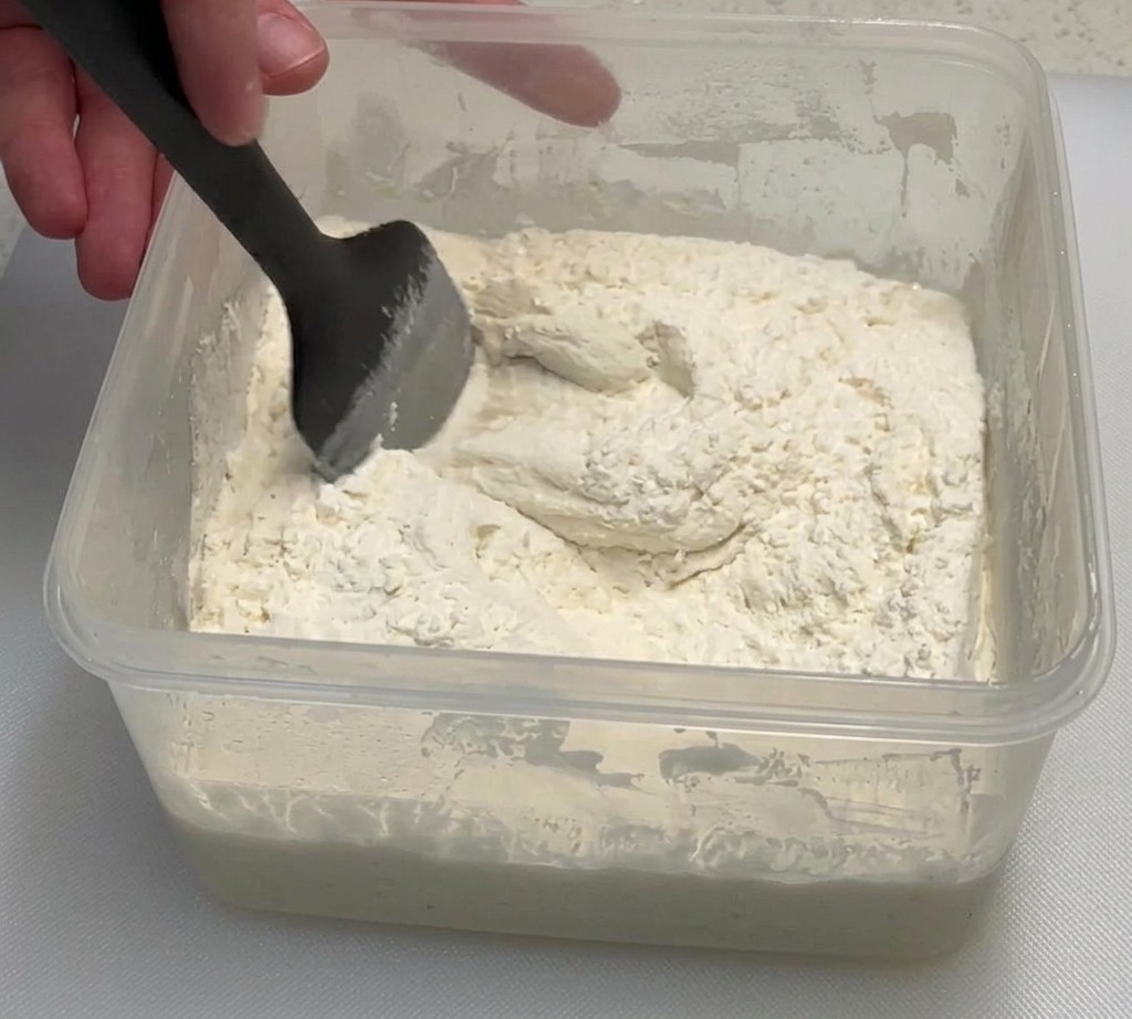 A hand using a black spatula to mix a sticky flour and poolish mixture in a clear plastic container.