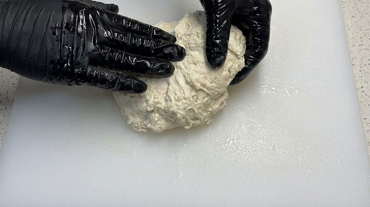 Hands wearing black gloves kneading a wet, shaggy pizza dough on a white cutting board.