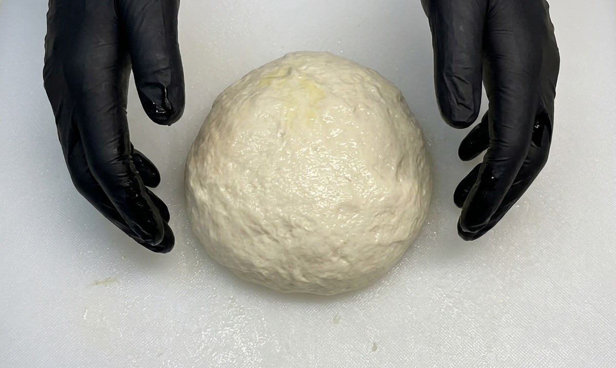 Black-gloved hands tucking and shaping a smooth, round ball of pizza dough on a white board.