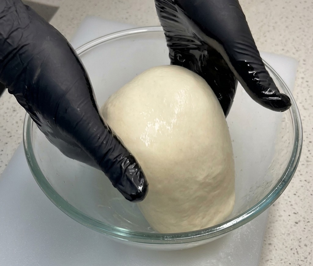 Gloved hands lifting and stretching the center of a soft pizza dough upwards inside a glass mixing bowl.