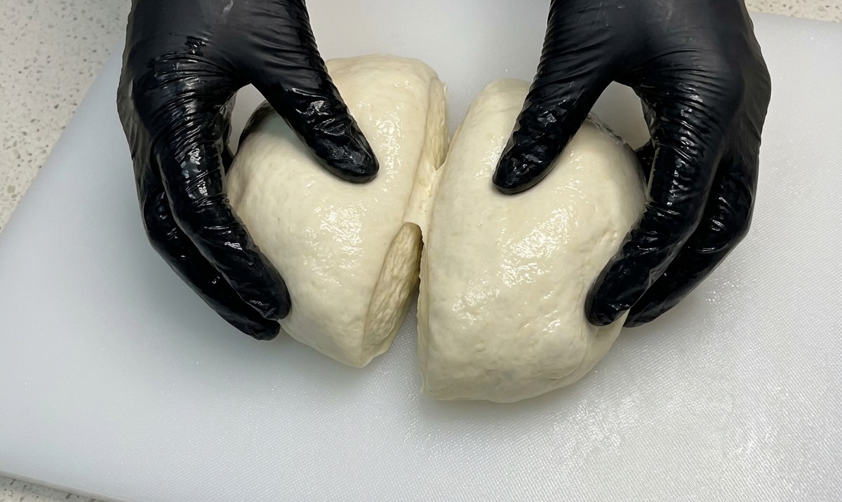 Hands in black gloves pulling apart a large, airy dough ball into two equal halves on a white cutting board.