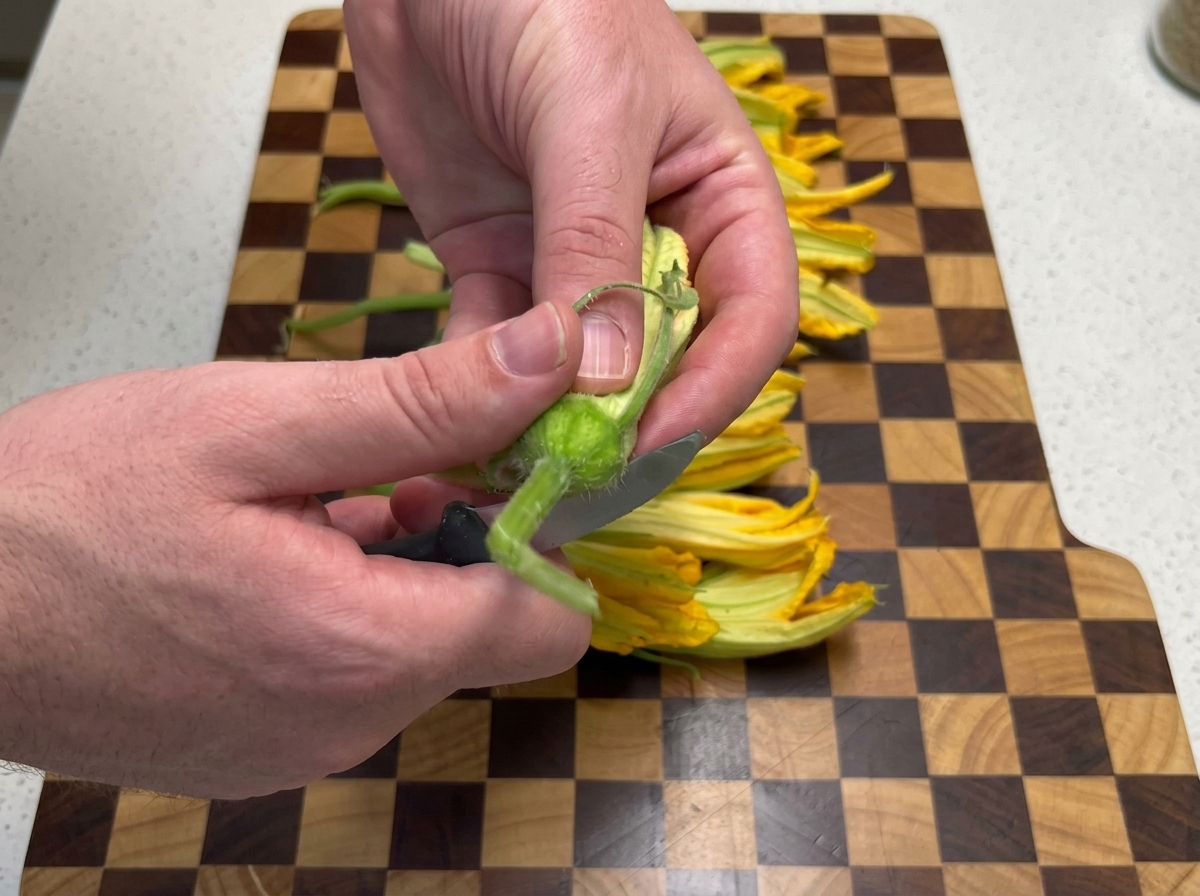 Hands using a small paring knife to trim the stem from a fresh yellow pumpkin flower on a wooden checkerboard cutting board.