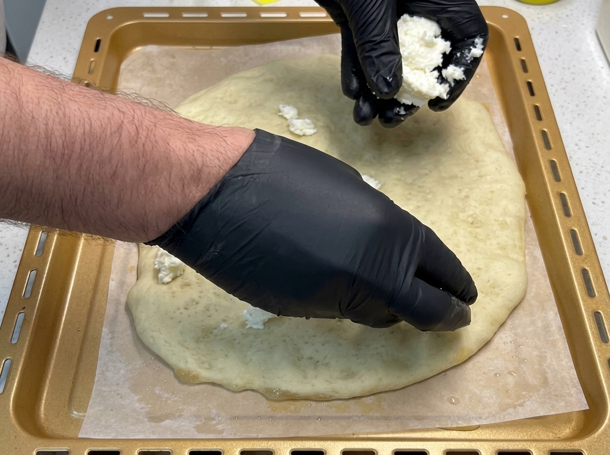 Black-gloved hands placing torn chunks of white fresh mozzarella cheese onto a golden, par-baked pizza dough on a baking sheet.