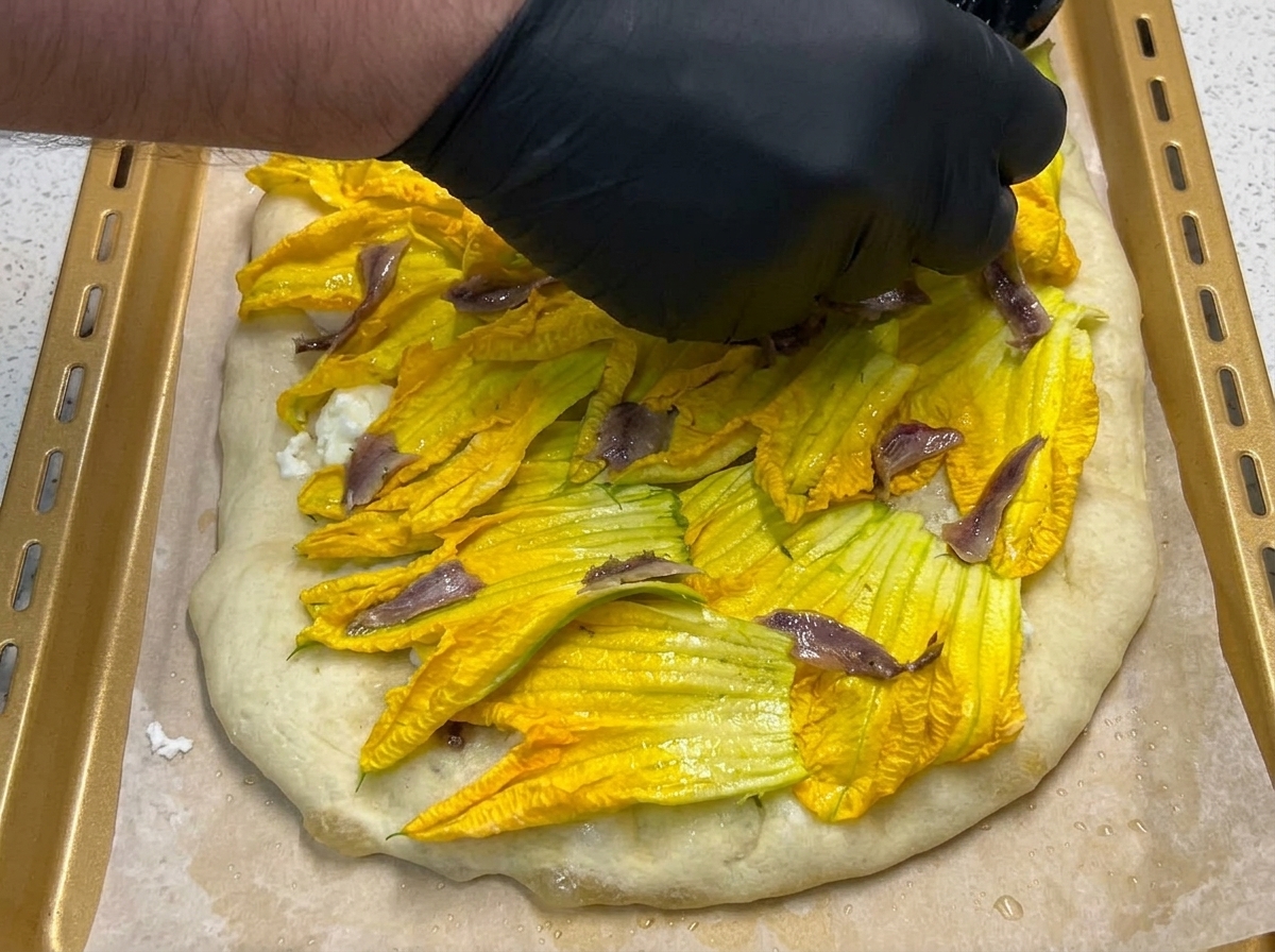 A black-gloved hand arranging small, dark anchovy fillets on top of vibrant yellow pumpkin flowers on a pizza.