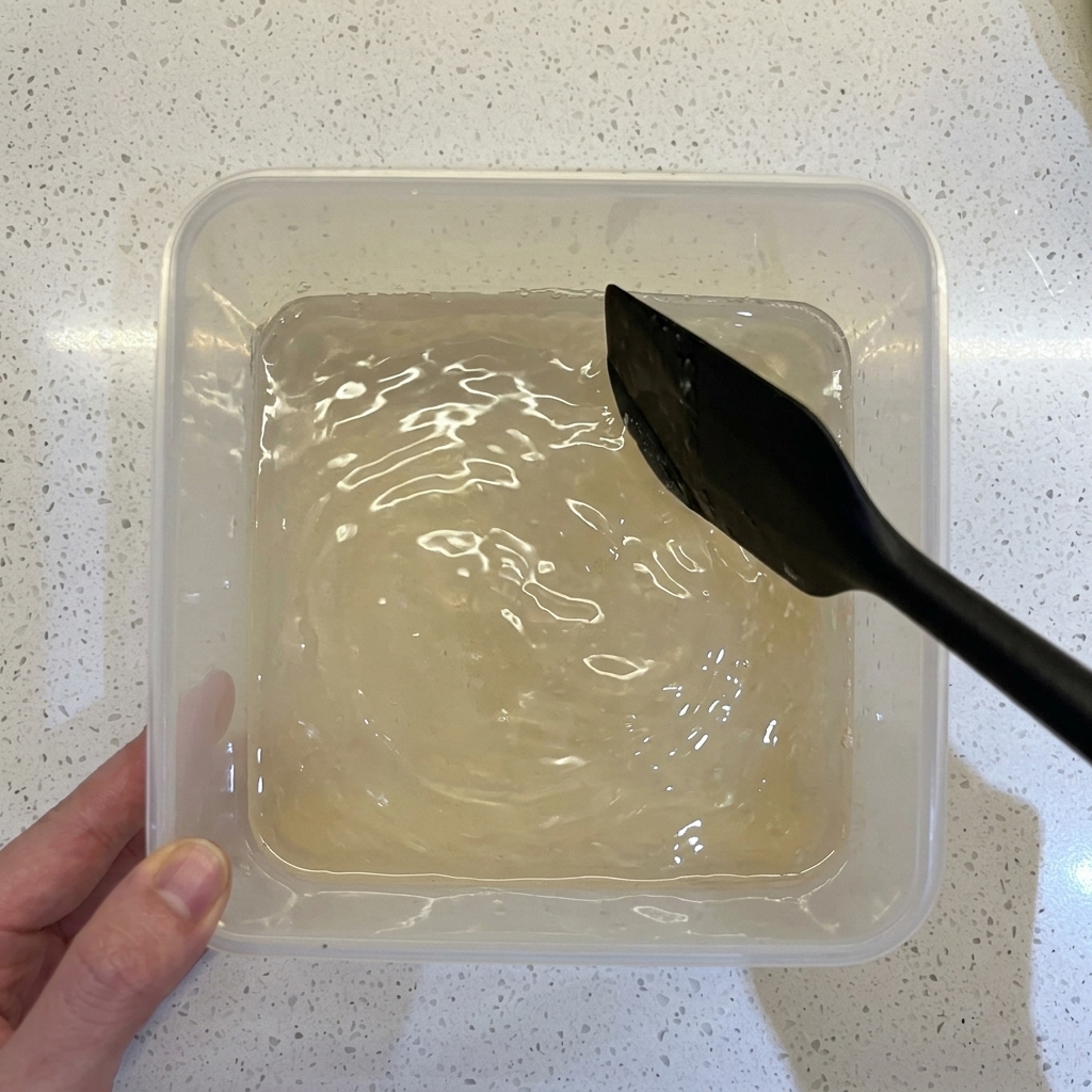 A black spatula stirring a watery flour and yeast mixture inside a clear square plastic container.