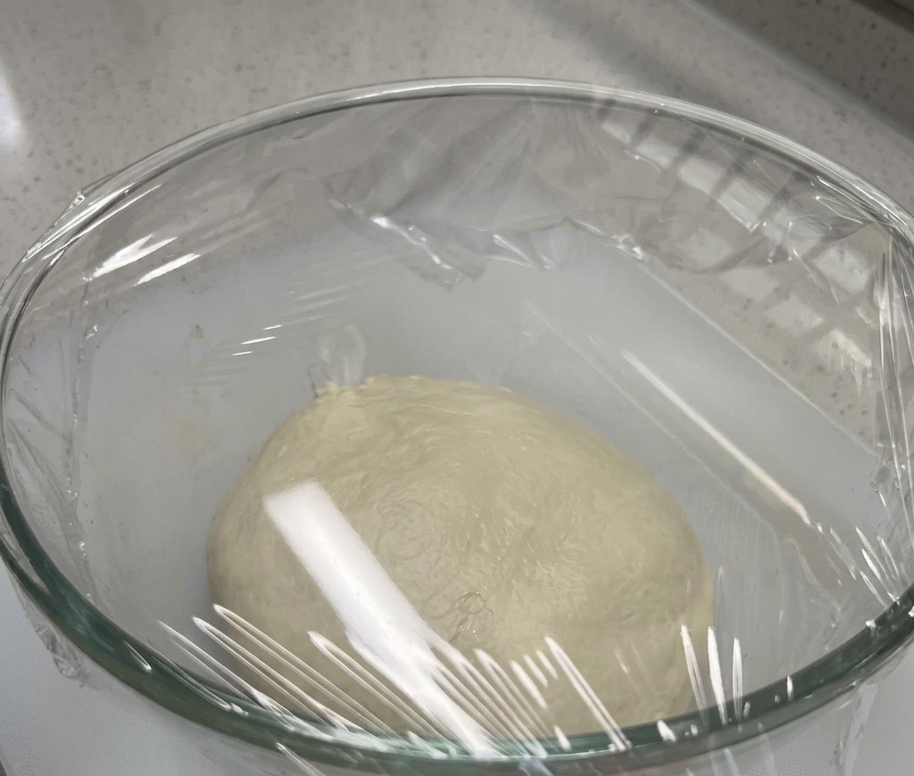 A smooth ball of pizza dough resting at the bottom of a large glass bowl, sealed tightly with plastic wrap.