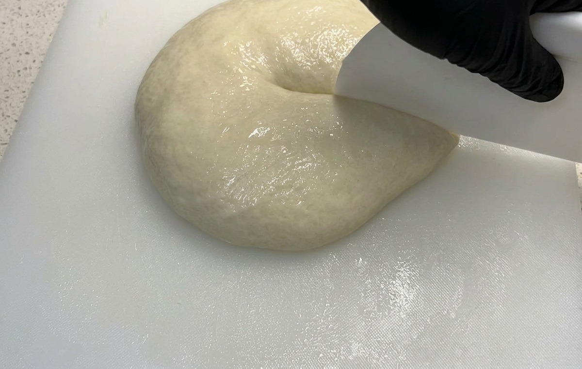A white bench scraper pressing down into a soft mound of pizza dough to divide it in half.