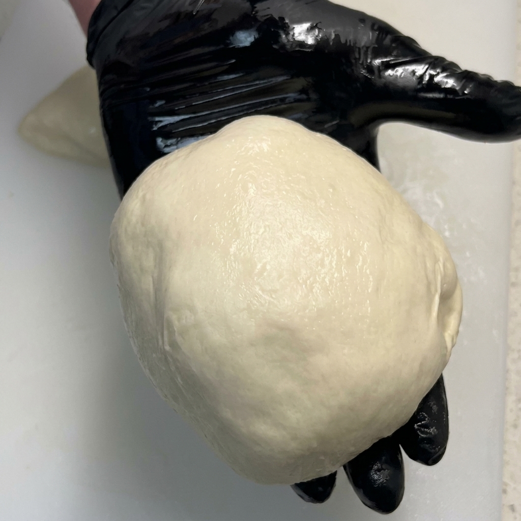 A shiny, perfectly smooth ball of pizza dough resting in the palm of a black-gloved hand.