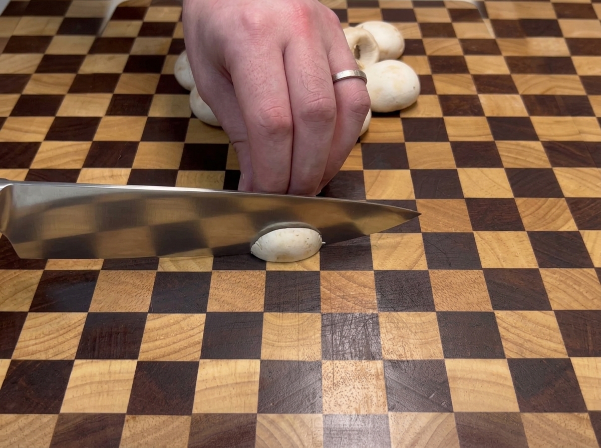 A hand using a large chefs knife to slice white button mushrooms on a two-toned checkered wooden cutting board.