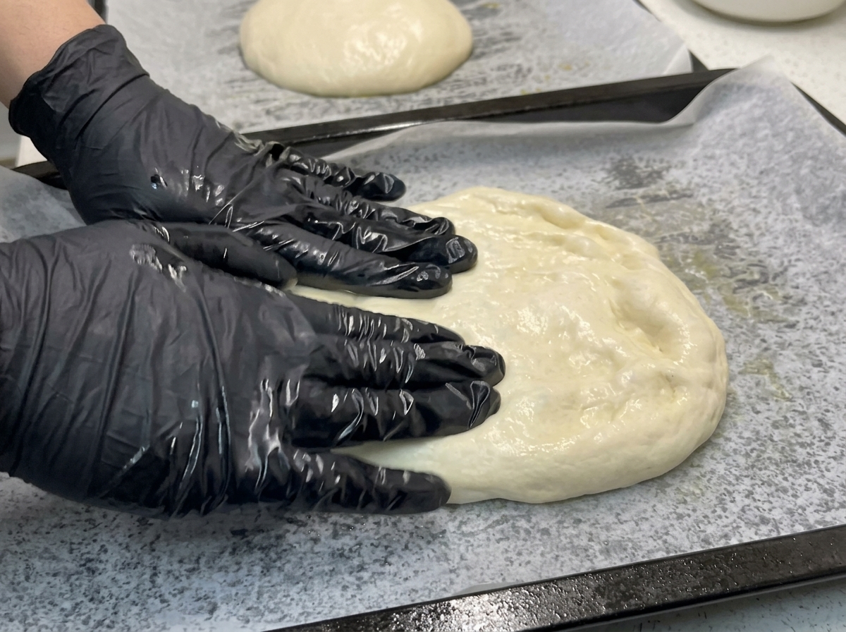 Gloved hands gently pressing and stretching a soft mound of pizza dough flat onto a parchment-lined baking sheet.