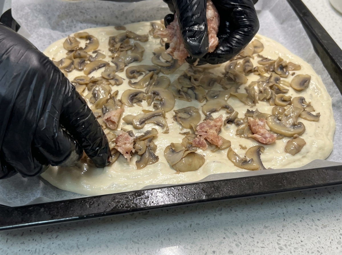 Gloved hands dropping pieces of raw sausage meat onto pizza dough already topped with sautéed mushrooms.