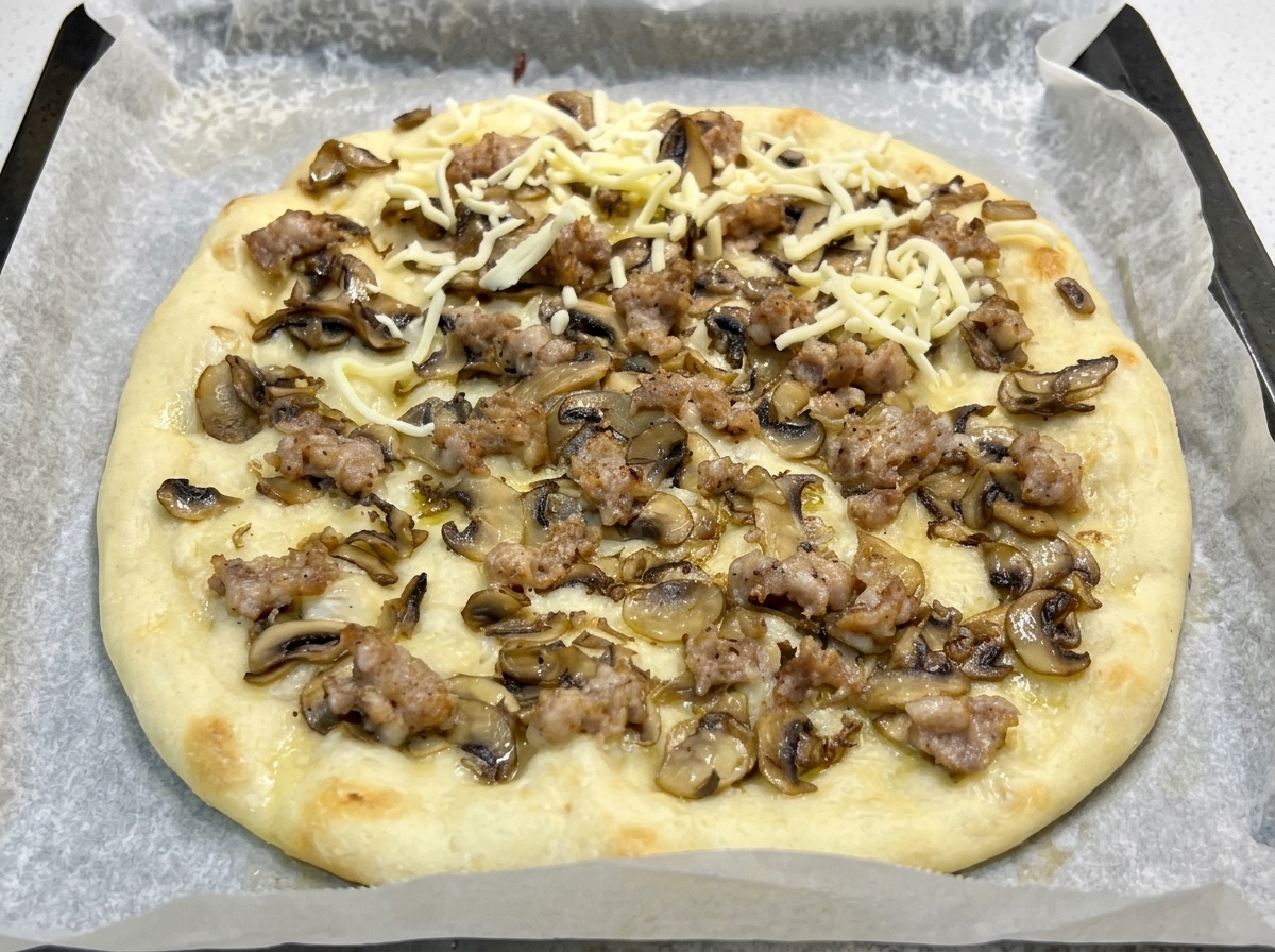 A par-baked pizza base on parchment paper topped with cooked mushrooms, sausage, and scattered shreds of mozzarella cheese.