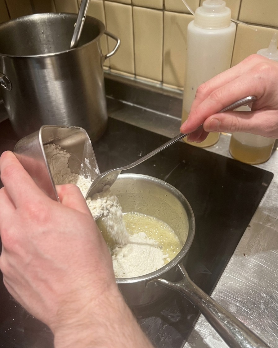 Pouring a scoop of flour into melted butter in a saucepan to make a roux.