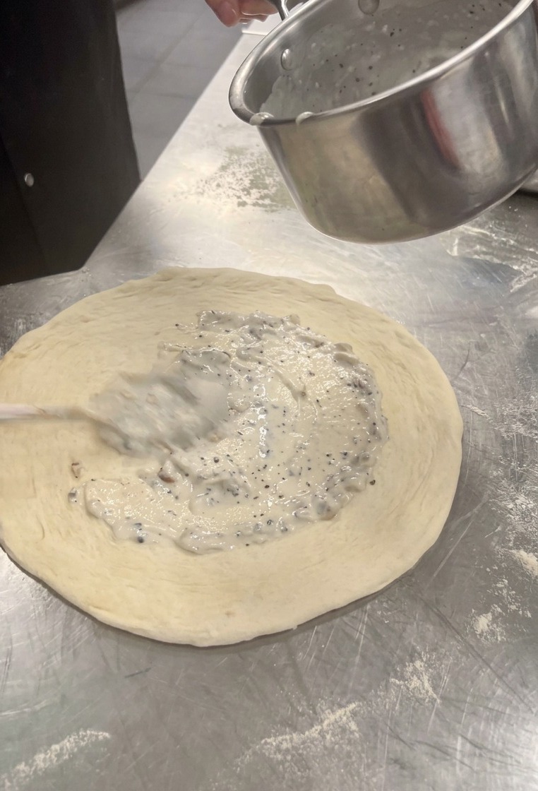 Spreading creamy white mushroom sauce over the center of the stretched pizza dough using a spoon.