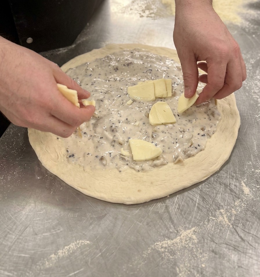 Hands placing small pieces of mozzarella cheese evenly over the white sauce on the raw pizza dough.