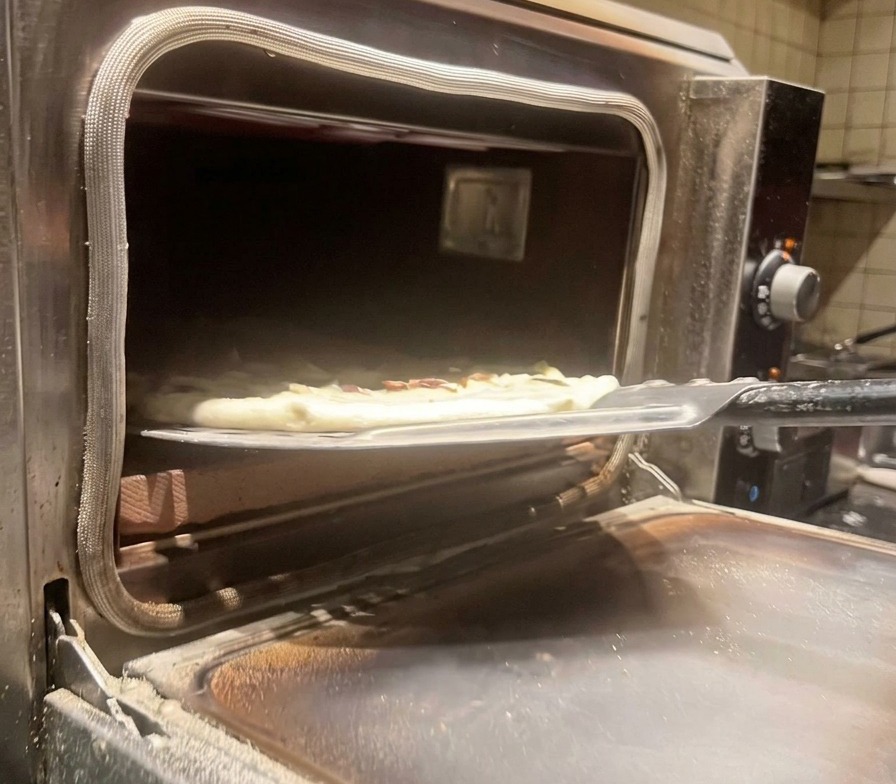 A raw mushroom pizza being transferred into a hot commercial pizza oven using a metal pizza peel.