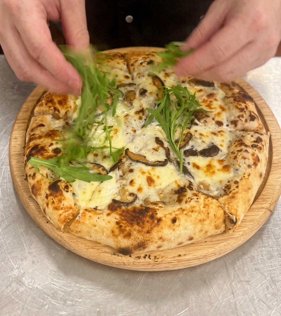 Hands arranging vibrant green arugula leaves on top of a baked and sliced mushroom pizza resting on a wooden board.