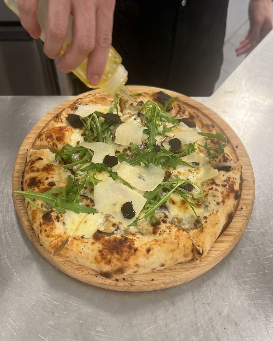 A hand squeezing a clear bottle to drizzle golden truffle oil over a finished mushroom, arugula, and truffle pizza.