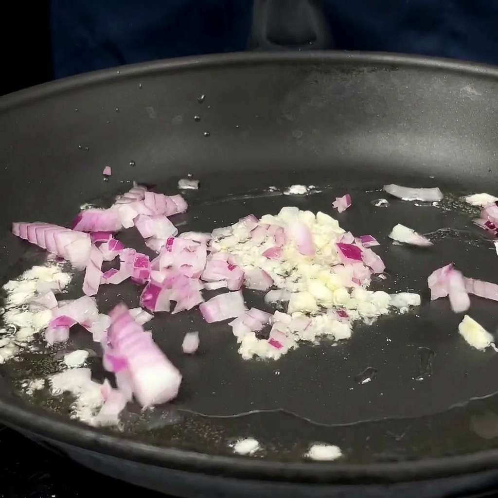 Diced red onions and minced garlic sizzling in hot oil in a dark non-stick skillet.