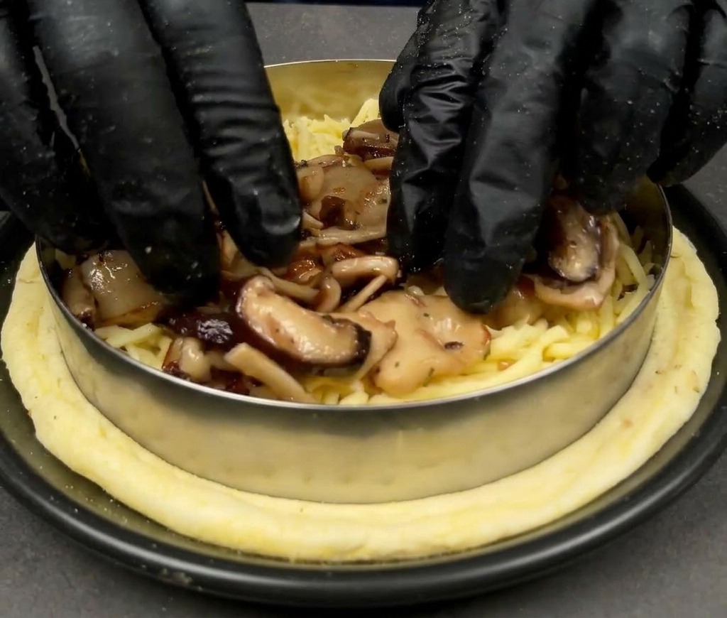 Hands wearing black gloves placing cooked mixed mushrooms over a bed of mozzarella cheese on a pizza base.