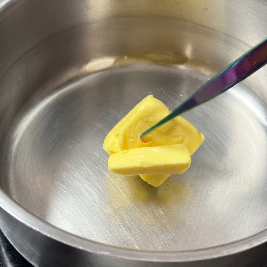 A square block of yellow butter melting in a shiny stainless steel pot.