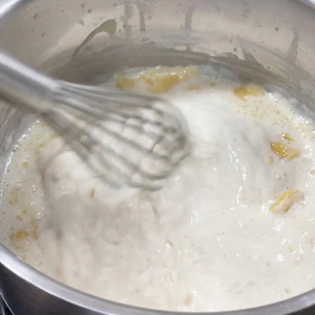 A whisk vigorously stirring a thick, bubbling white sauce in a stainless steel pot.