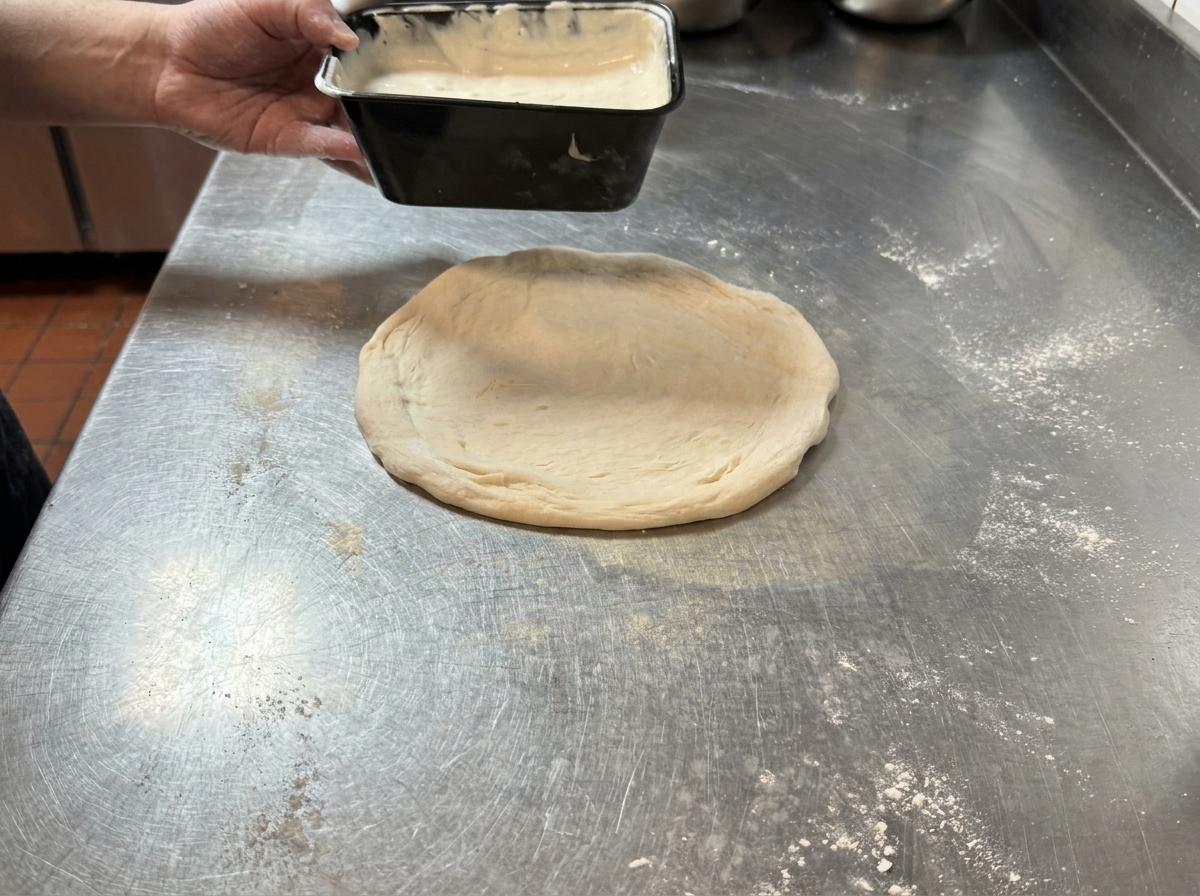 A hand pouring creamy black truffle white sauce from a container onto a round piece of stretched raw pizza dough resting on a stainless steel table.