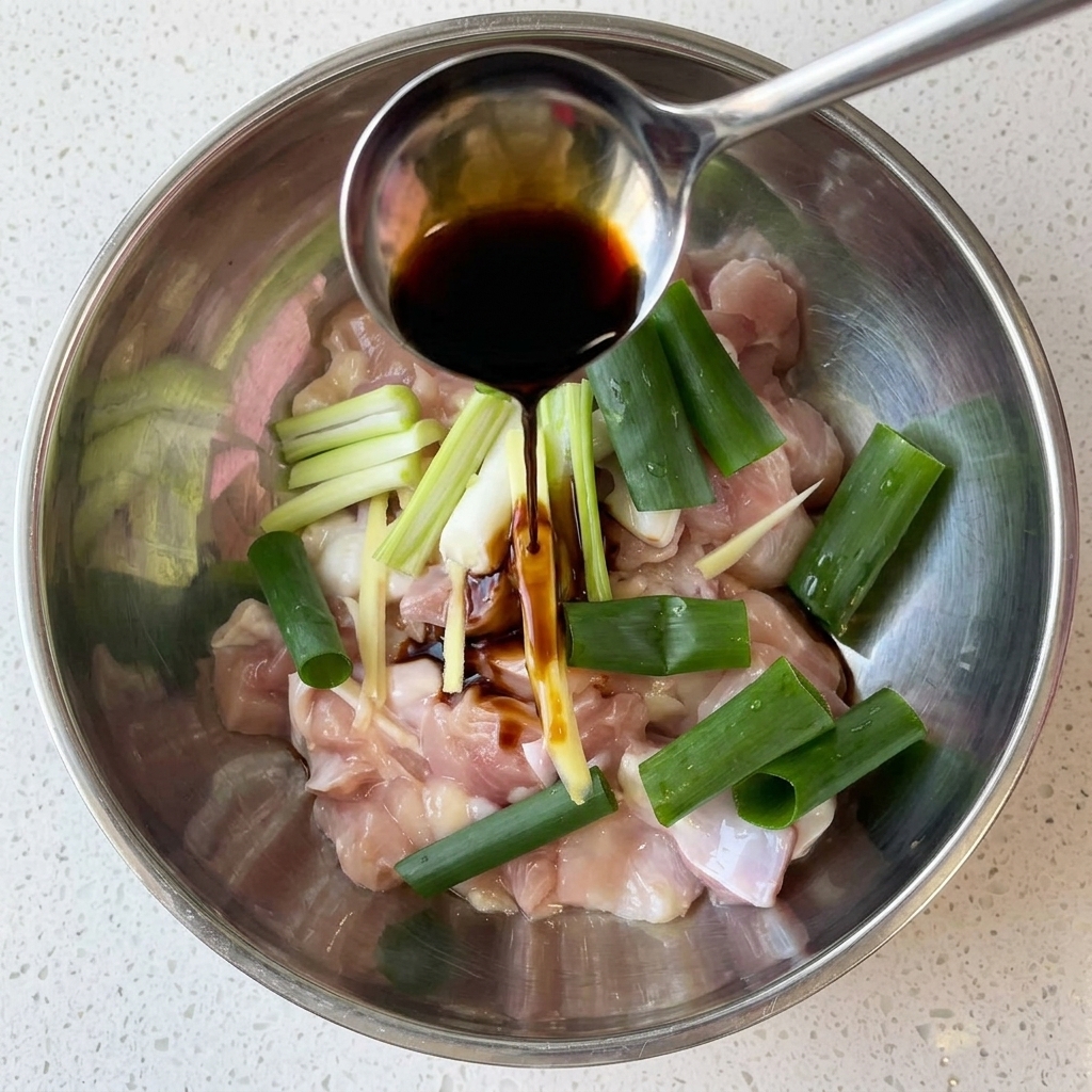 Raw chicken chunks in a metal mixing bowl being drizzled with dark soy sauce alongside green onions and ginger.