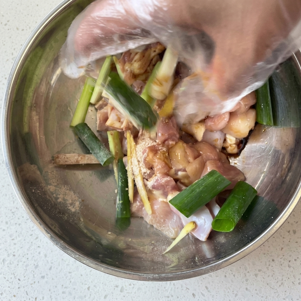A hand wearing a plastic glove mixing raw chicken pieces with green onions, ginger, and marinade in a metal bowl.