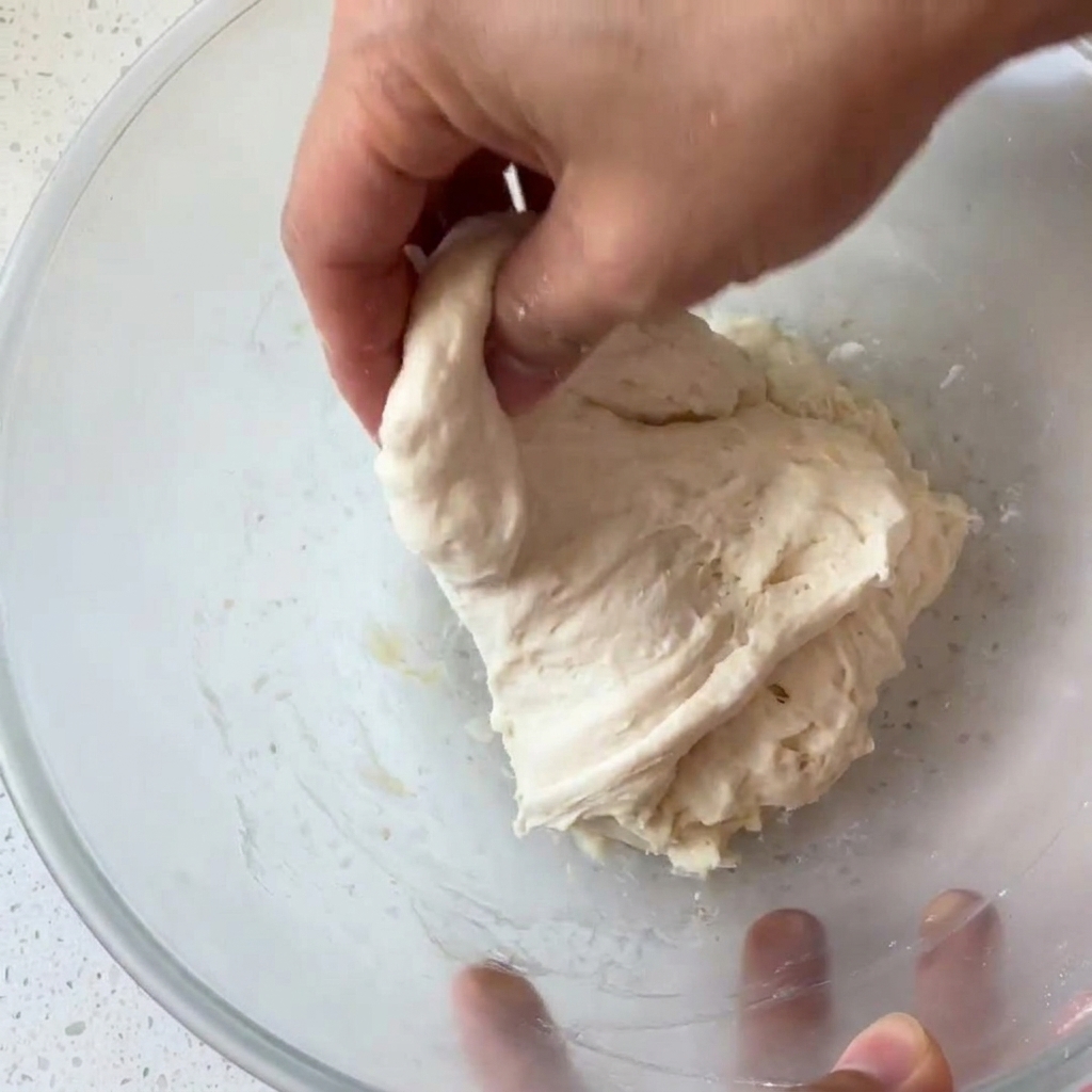 A bare hand grabbing and folding a wet, sticky ball of dough inside a glass bowl.