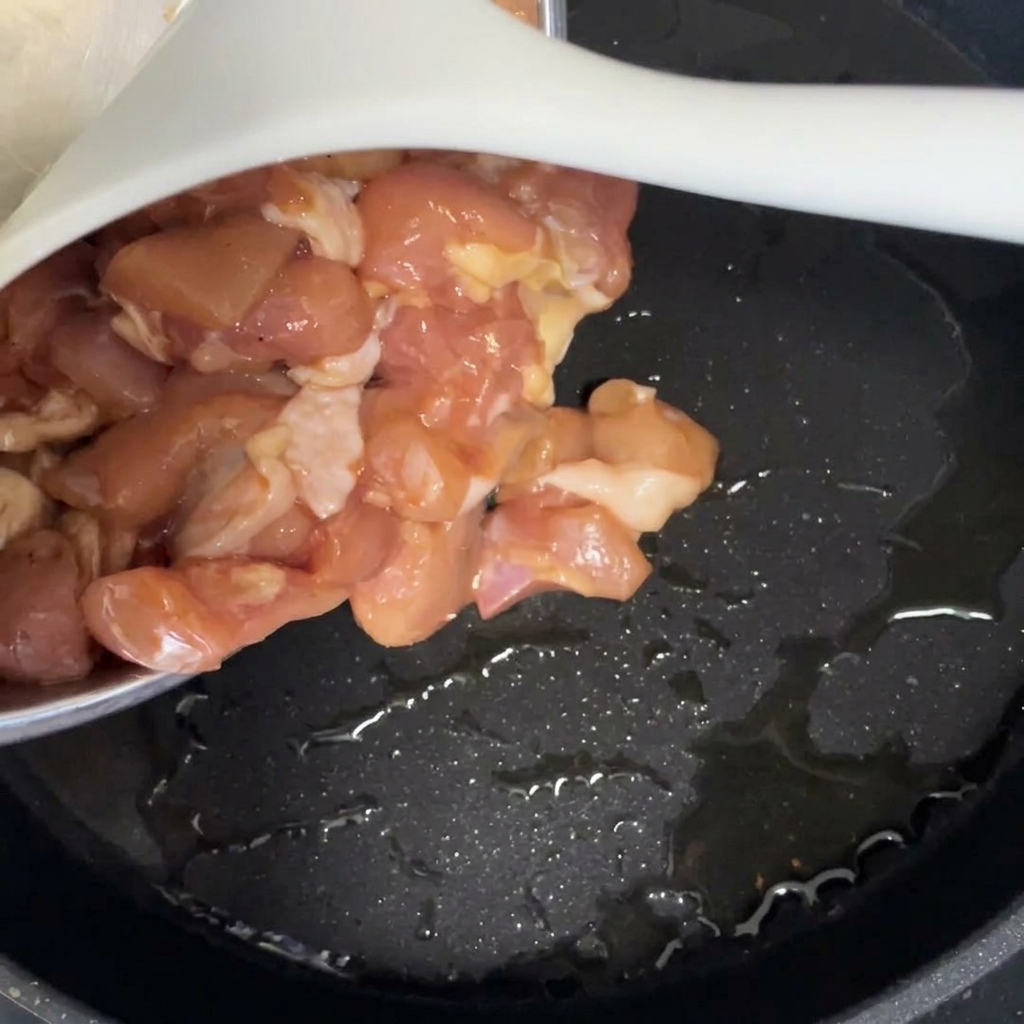 Raw marinated chicken pieces being poured into a black skillet coated with hot cooking oil.
