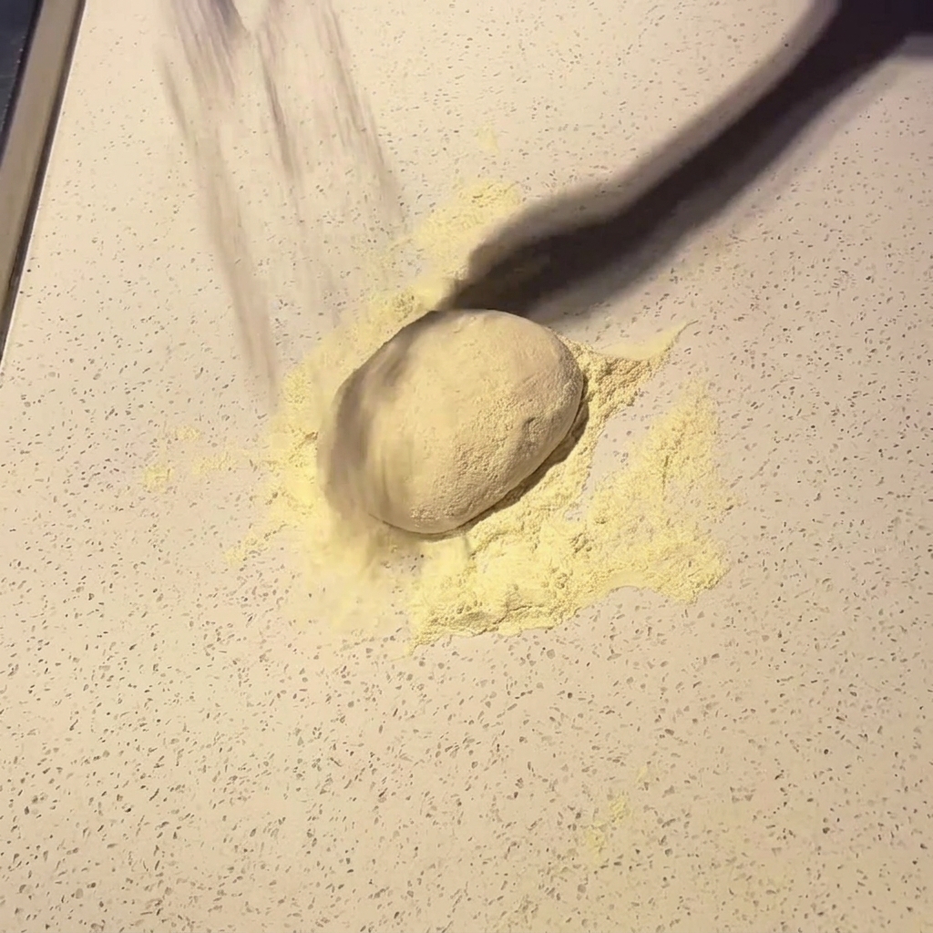 A ball of pizza dough being dusted with flour on a light countertop.
