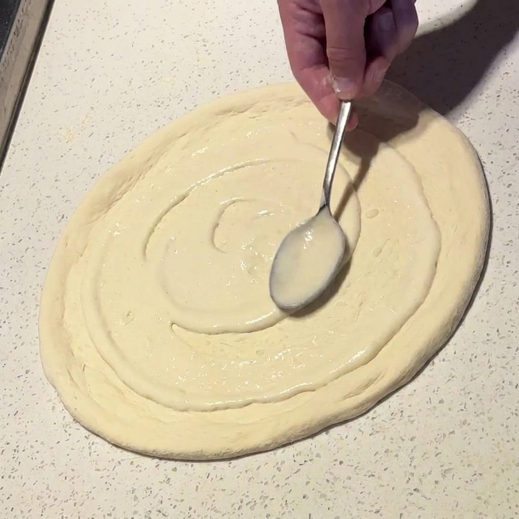 A metal spoon spreading creamy white sauce in a circular motion over a pizza dough base.