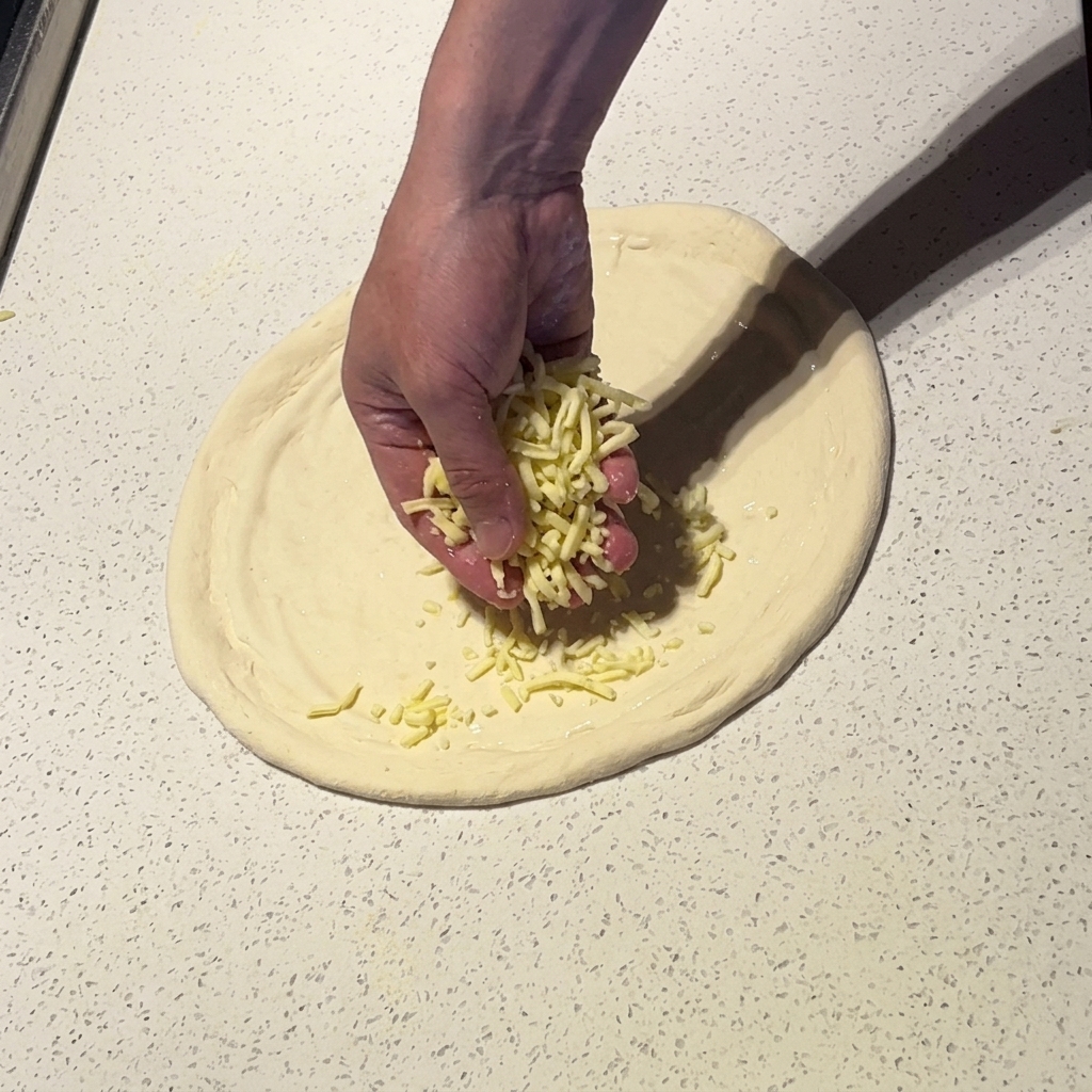 A hand sprinkling shredded mozzarella cheese over stretched pizza dough coated in white sauce.