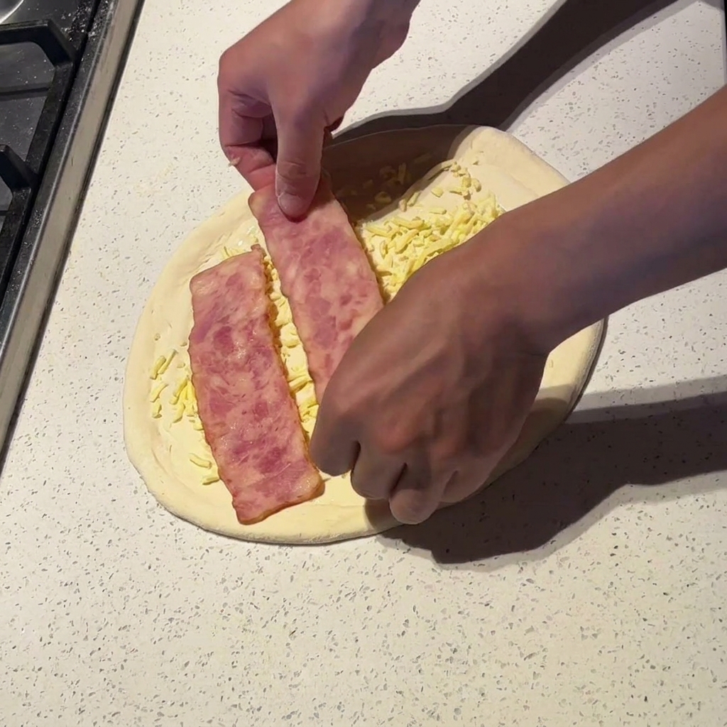 Hands placing wide strips of lightly cooked bacon onto a pizza topped with white sauce and cheese.