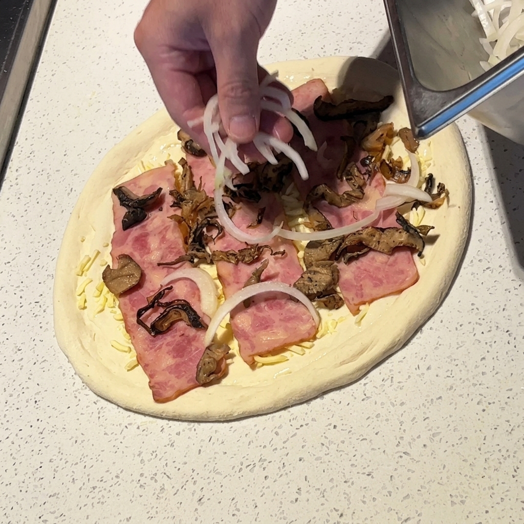 A hand scattering thin slices of white onion onto a pizza topped with bacon and mushrooms.