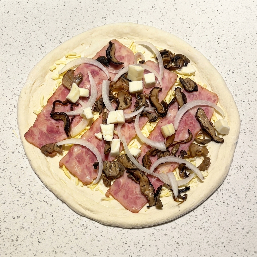 A fully assembled raw pizza topped with bacon, mushrooms, onions, and small cubes of fresh mozzarella.