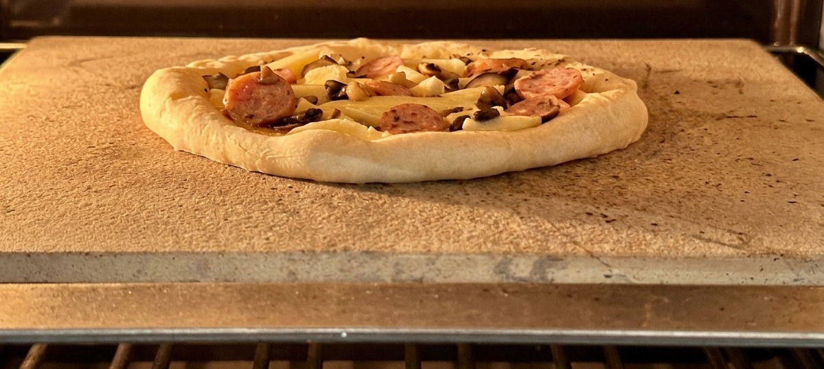 An assembled pizza with sausage and mushrooms sitting on a baking stone inside a hot oven.