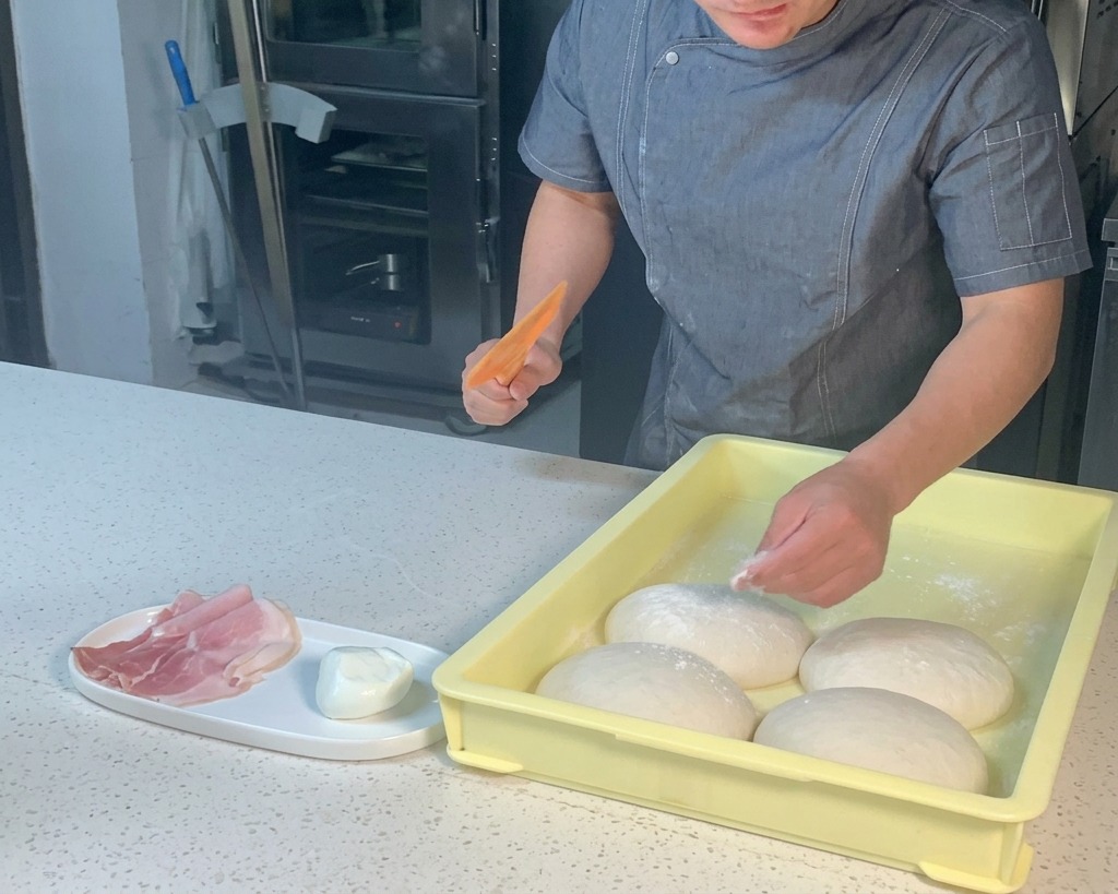 Chef using an orange plastic scraper to lift a ball of pizza dough from a yellow tray.