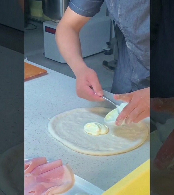 Chef using the back of a spoon to evenly spread a rich white sauce onto a stretched pizza dough in a circular motion.