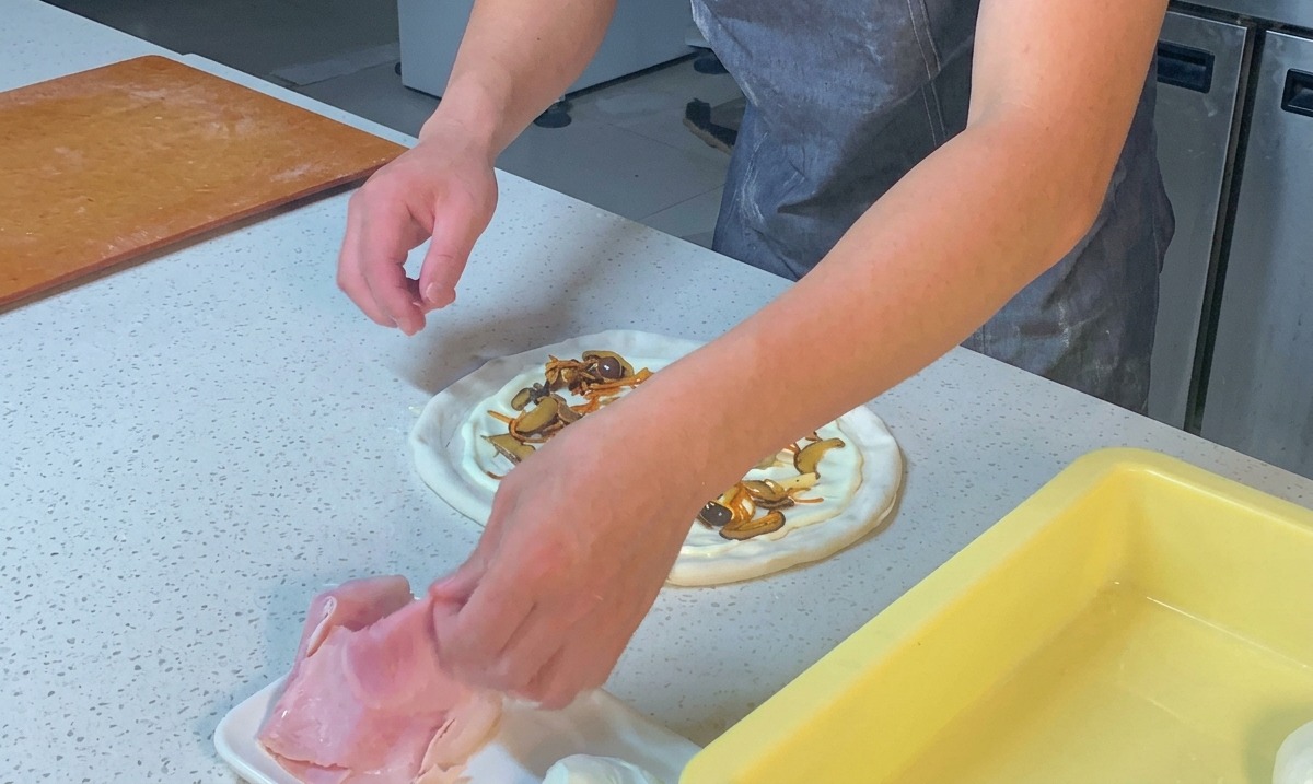 Chefs hands tearing slices of pink ham and scattering the pieces onto a pizza crust topped with white sauce and mushrooms.