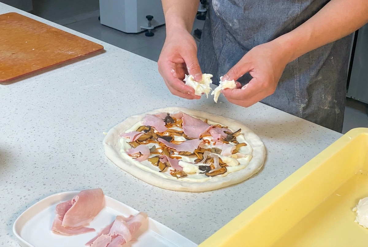 Chefs hands tearing fresh white mozzarella cheese over a pizza topped with ham, mushrooms, and cordyceps.