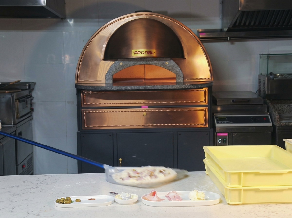 A raw ham and mushroom pizza being swiftly moved on a long-handled metal peel toward a glowing dome oven.